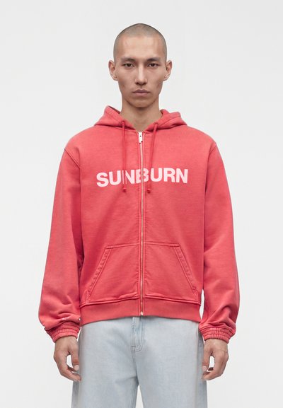Homme à la tête rasée portant un sweat à capuche rouge zippé avec le texte « SUNBURN » et un jean bleu clair, debout devant un fond blanc uni.