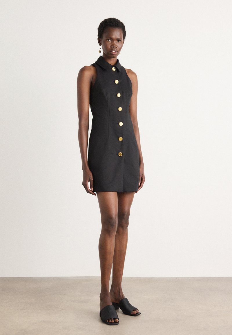 Črna brezrokavna obleka s kragnim izrezom, ki ima zlate gumbe spredaj. Obuta z črnimi natikači. Gladka tekstura.