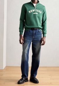 Zelená mikina na zip s nápisem "NEW YORK", doplněná modrými džíny. Boty jsou černé, z texturované kůže s masivní podrážkou.