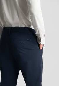 Sötétkék pamut nadrág, sima textúrával. Hátsó zseb gombbal és finom varrási részletekkel.