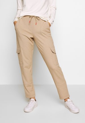 Pantalons cargo beiges avec une taille à cordon, deux poches latérales et des poignets côtelés. Fabriqués en un tissu doux et texturé. Stylés avec des baskets blanches.