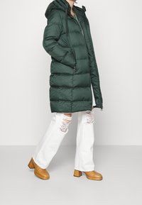 Gevoerde groene parka met een capuchon, voorzien van een rits aan de voorkant, zijzakken en een textuurstof. G gedragen met witte gescheurde jeans en bruine plateauboots.