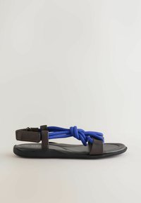 Sandale plate noire avec des lanières en cuir marron et un détail en corde nouée bleue au niveau du cou-de-pied, et une bride de cheville ajustable avec fermeture velcro.