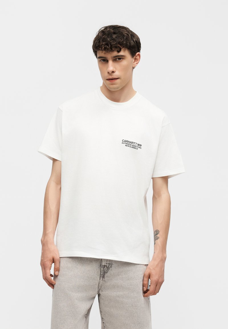 Giovane uomo con capelli corti e ricci che indossa una t-shirt bianca Carhartt WIP e jeans grigio chiaro, in piedi davanti a uno sfondo bianco uniforme.