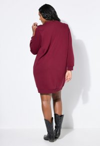 Mujer vestida con un vestido burdeos de manga larga y botas de vaquero negras, de pie y de espaldas contra un fondo blanco liso.