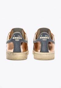 Le sneakers Diadora presentano una parte superiore in suede grigio con accenti metallici in rame, una suola beige testurizzata e un dettaglio del logo cucito sulla punta.