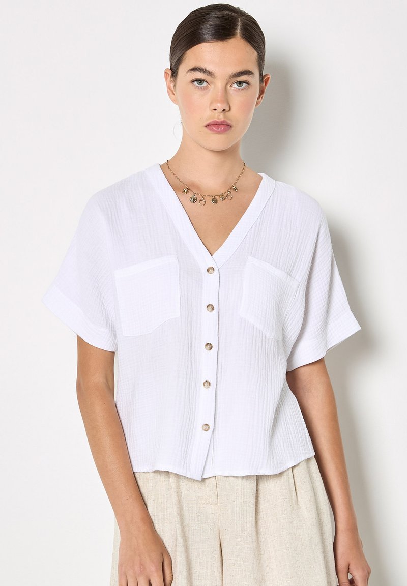Femme aux cheveux foncés portant un chemisier blanc à manches courtes boutonné avec deux poches poitrine et un pantalon beige, portant un collier à breloques en or.