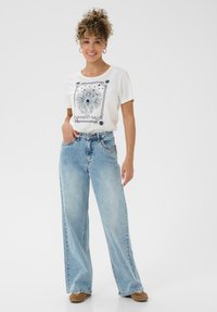 Hvid bomulds-t-shirt med et grafisk tryk af en løve og stjerner. Kombineret med lyseblå, højtaljede jeans med vide ben. Brune sko.