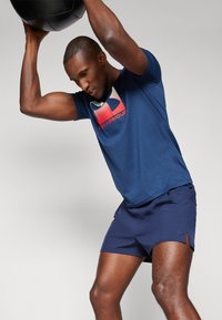 Marineblå atletisk skjorte med et rødt og hvidt logo, parret med marineblå shorts med sideskæringer. Mand løfter en sort vægtet bold over hovedet.