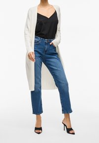 Kvinna i blå högmidjade jeans, svart camisole, lång vit cardigan och svarta öppna klacktofflor, står mot vit bakgrund.