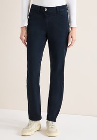Femme portant un pantalon bleu foncé à jambes droites avec des poches avant et des baskets blanc cassé sur un fond neutre.