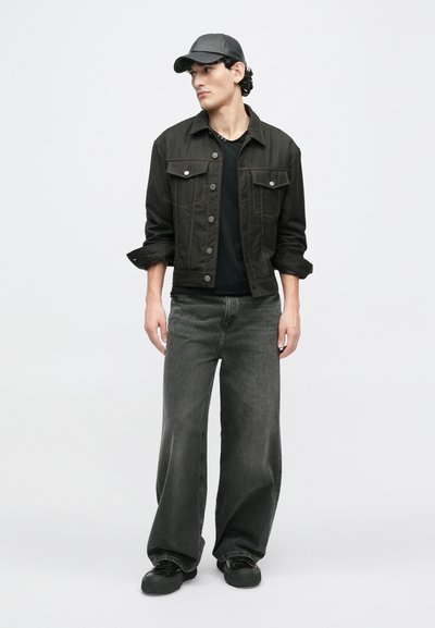 Jachetă din denim negru cu nasturi, mâneci lungi și detalii la buzunare, purtată cu blugi negri cu croială largă și o șapcă neagră. Textură netedă pe întreaga suprafață.