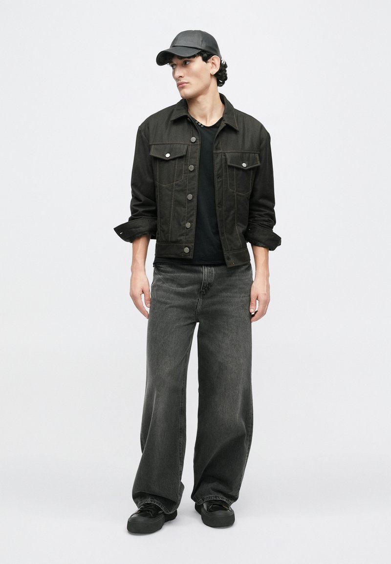 Sort denimjakke med knapper, lange ærmer og lomme-detaljer, parret med vide sorte jeans og en sort kasket. Glat tekstur hele vejen igennem.