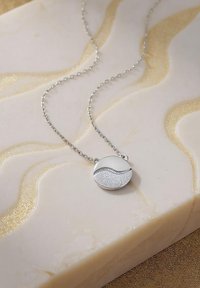 Silverhalsband med en rund hänge som har en slät, polerad halva och en texturerad, glittrig halva, placerad mot en ljus marmorbakgrund.