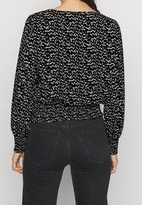 Schwarz-weiße gemusterte Bluse mit langen Ärmeln, elastischem Bund und eng anliegenden Manschetten, kombiniert mit schwarzen Jeans. Strukturiertes, lässiges Design.