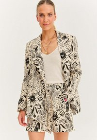 Beige blazer a šortky s černými abstraktními vzory. Texturovaná tkanina s klopovým límcem. Nosič přes krémový top.