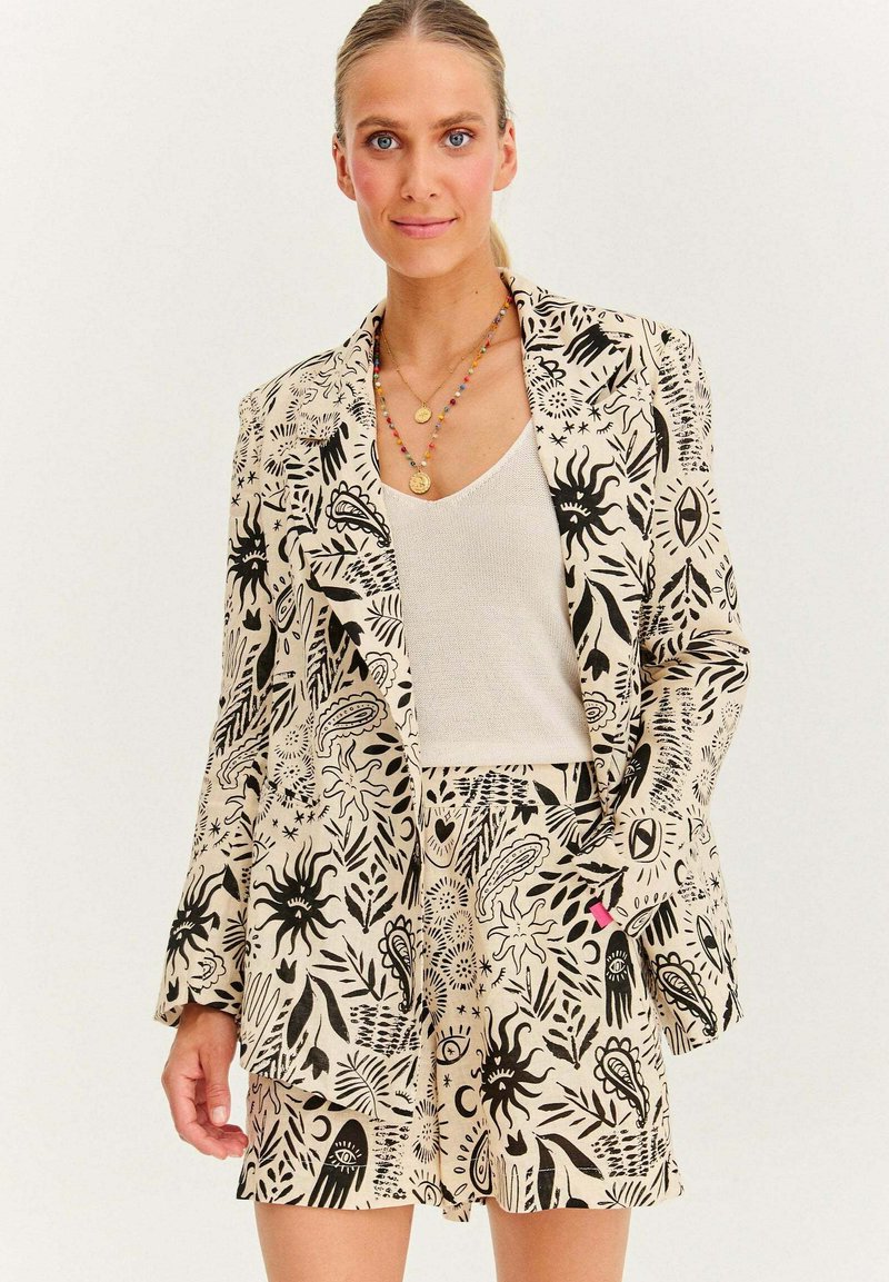 Beige blazer a šortky s černými abstraktními vzory. Texturovaná tkanina s klopovým límcem. Nosič přes krémový top.