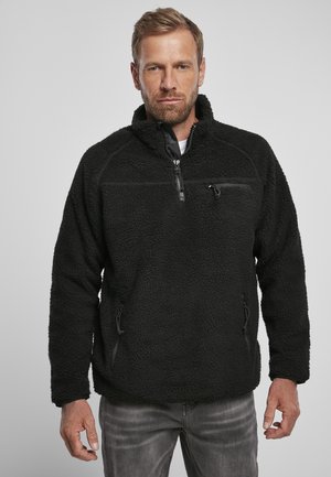 Pull en polaire noir avec col zippé, extérieur texturé et deux poches latérales. Comprend une poche poitrine zippée et des cordons élastiques.