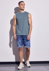 Maglietta senza maniche teal da uomo, pantaloni shorts in denim con risvolti e sneakers chiare. Design semplice, texture morbida e vestibilità casual.