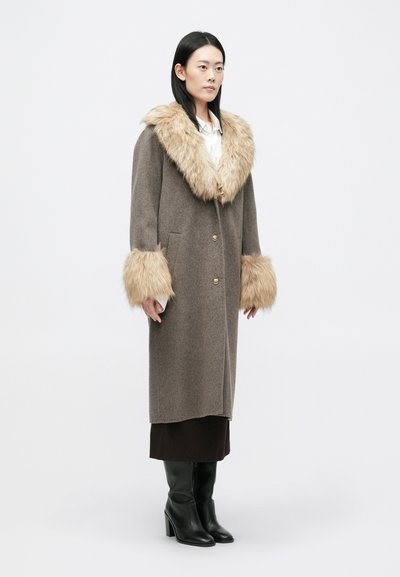 Καφέ μάλλινο παλτό �με ψεύτικη γούνα στον γιακά και τα μανίκια, διακοσμημένο με χρυσά κουμπιά και πλαϊνές τσέπες, φορεμένο πάνω από μια σκούρα φούστα και μαύρες μπότες με τακούνι.