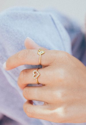 Deux bagues en or sur une main ; l'une présente un contour de cœur serti de pierres claires, l'autre a un cœur en or avec une gemme colorée.