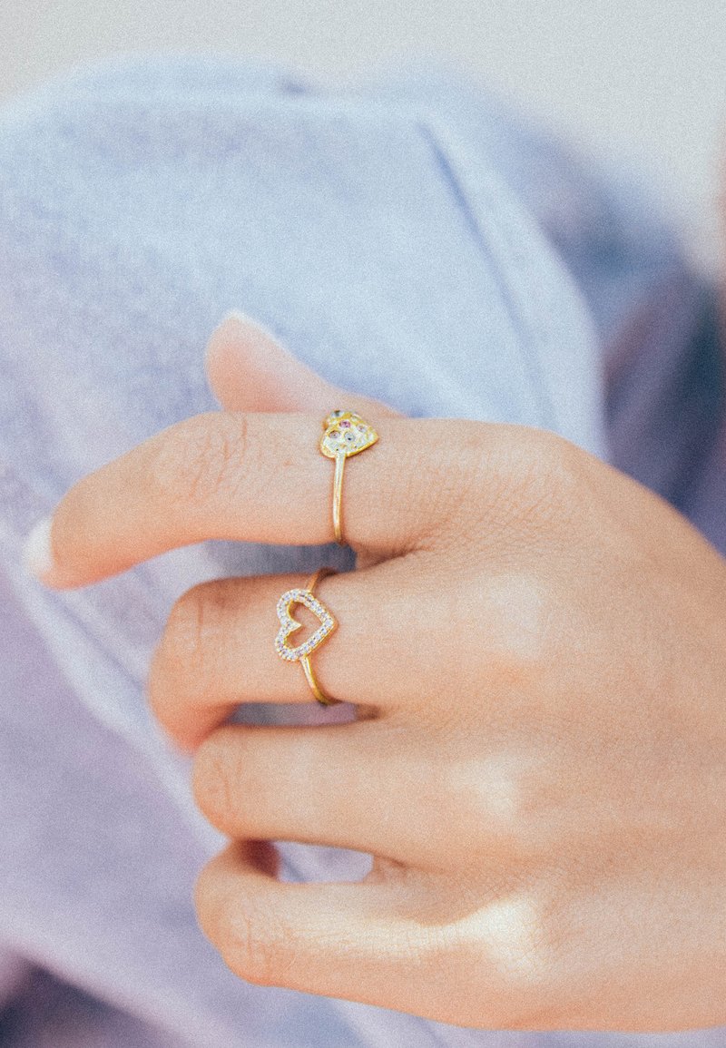 Twee gouden ringen op een hand; de ene heeft een hartomtrek bezet met heldere stenen, de andere heeft een gouden hart met een kleurrijke edelsteen.