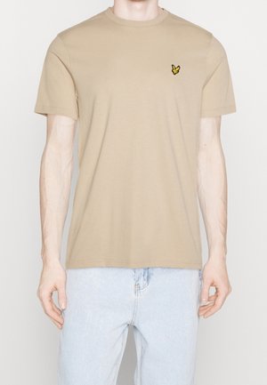 Mann trägt ein beiges T-Shirt mit kurzen Ärmeln und einem kleinen gelb-schwarzen Vogel-Logo auf der linken Brust, kombiniert mit hellblauen Jeans.