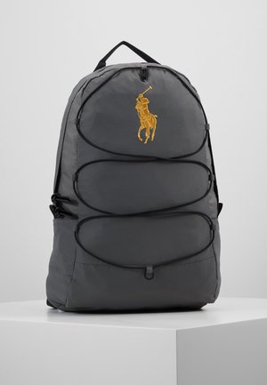 Tmavě šedý batoh s třemi předními kapsami na zip a žlutým vyšitým logem hráče polo na horní kapse, stojící na bílém povrchu.