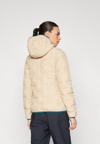 Beige dunjakke med hette, med kviltet tekstur og en kontrastfarge i grønnn nederkant. Bakvinduet viser en strømlinjeformet, tettsittende design.