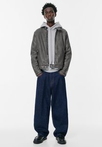 Giovane uomo in piedi con le mani in tasca, che indossa una felpa grigia, una giacca di pelle grigia scura, pantaloni larghi blu scuro e scarpe nere.