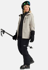 Tenue de ski avec une veste imperméable gris clair, un pantalon isolant noir, des gants noirs, un casque et des bâtons de ski. Présente des logos Jack Wolfskin.