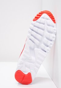 Semelle de sneaker Nike blanche et rouge inclinée contre un mur blanc, montrant un motif de grip texturé et le logo de la marque sur la semelle.