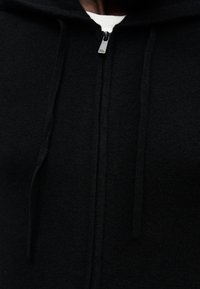 Sweat à capuche zippé noir en tissu doux, avec poche kangourou, capuche à cordon de serrage et détail de fermeture éclair argentée.