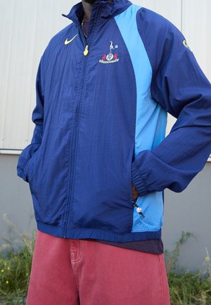 Une personne porte une veste de Tottenham Hotspur bleu marine et bleu clair ainsi qu’un pantalon rouge, et se tient debout les mains dans les poches.