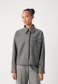 Black and white checkered cropped jacket with a buttoned collar, single chest pocket, and long sleeves, paired with gray trousers.