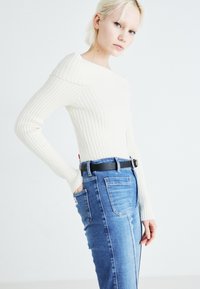 Top bianco a righe con maniche lunghe e design off-shoulder, abbinato a jeans in denim blu con una cintura nera e tasche posteriori.
