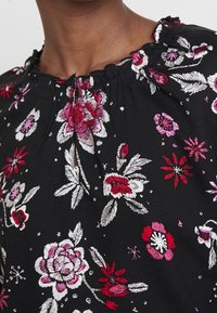 Haut en tissu noir avec broderies florales en rouge, rose et blanc. Présente un froncé au niveau de l'encolure et une petite patte de boutonnage à l'avant.