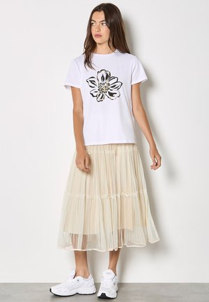 Młoda kobieta w białej koszulce z czarnym kwiatowym nadrukiem, beżowej plisowanej spódnicy midi z małymi kokardkami i białych tenisówkach, stojąca na tle białej ściany.