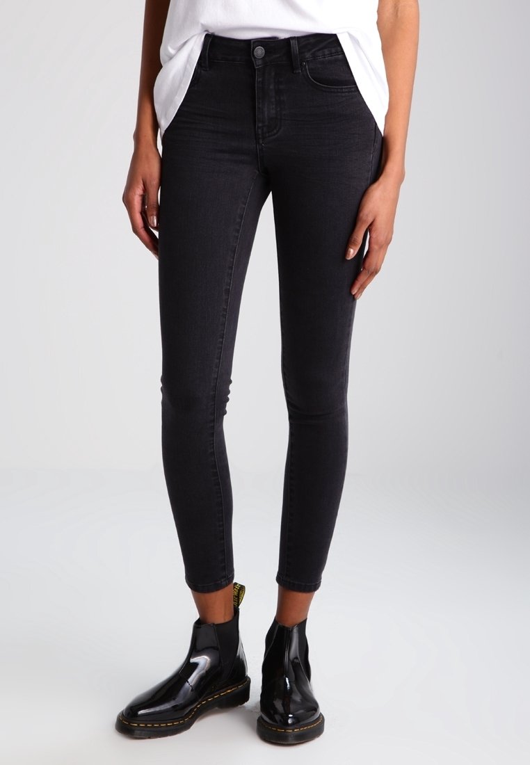 Personne portant un jean skinny noir et des bottines noires brillantes, debout devant un fond clair uni.
