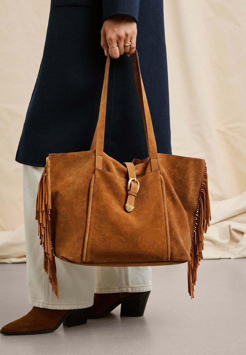 Borsa a spalla in suede marrone con dettagli in frange, manico in pelle e accessori in metallo. Forma robusta, portata a mano, abbinata a stivali con tacco.
