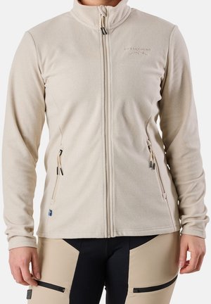 Beige fleecejack met volledige ritssluiting en twee ritssluiting zakken, gedragen over beige en zwarte ritssluiting broeken.