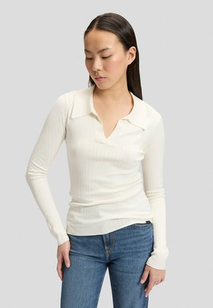 Kvinde med langt, glat hår har en tætsiddende cremefarvet ribbet langærmet top med krave og blå jeans, kigger ned med hænderne ved siderne.