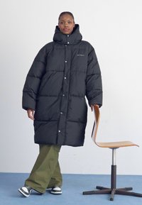 Zwarte pufferjas met een hoge kraag, grote gequiltte delen en drukknoopsluiting. Draag over losse groene broeken en gecombineerd met zwart-witte sneakers.