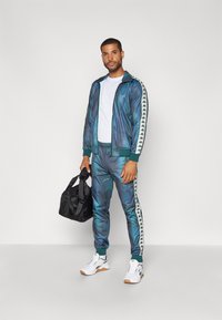 Chándal en degradado azul-verde con rayas laterales blancas que presentan logotipos. Combinado con una camiseta blanca y un bolso negro. Zapatillas blancas.