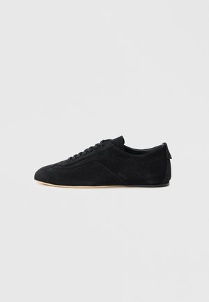Baskets basses en daim noir avec lacets et semelle beige, présentées de profil sur un fond gris uni.