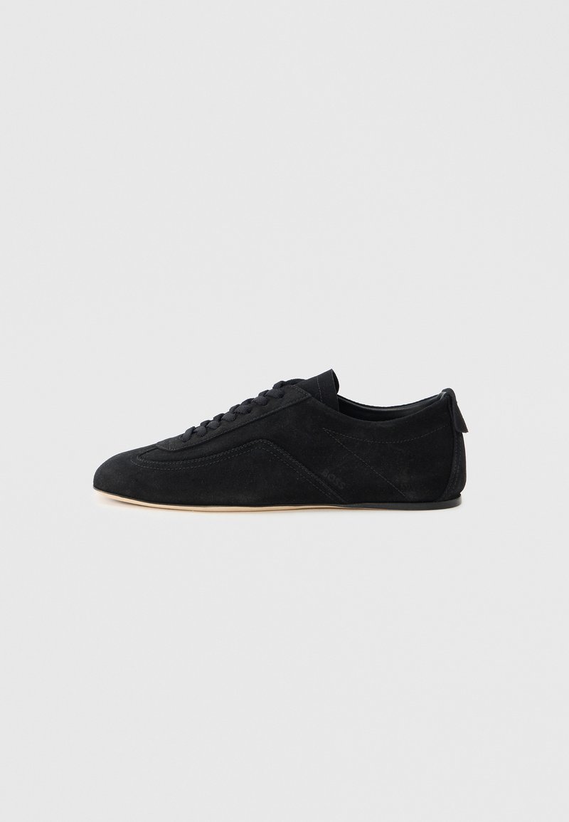 Baskets basses en daim noir avec lacets et semelle beige, présentées de profil sur un fond gris uni.