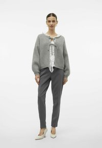 Maglione grigio over-sized con un nodo frontale, abbinato a pantaloni grigi su misura e décolleté color crema, con una blusa bianca a volant sotto.
