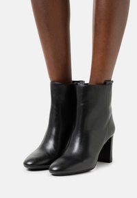 Botas de cuero negro hasta el tobillo con tacones cuadrados, llevadas sobre piernas de piel oscura, contra un fondo claro y liso.