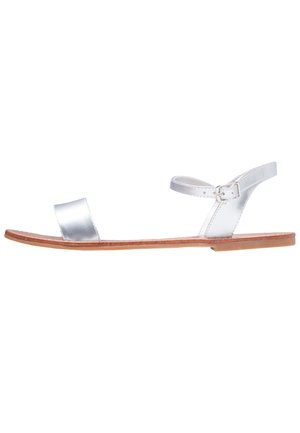 Srebrne metaliczne płaskie sandały z szerokim paskiem na palcach, paskiem wokół kostki z klamrą oraz brązową płaską podeszwą. Minimalistyczny design.