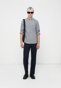 Hemd in hellgrauem Stoff, schlanke Passform, mit einer Brusttasche; kombiniert mit navyfarbenen Hosen und schwarzen Schuhen, accessoriert mit Sonnenbrille.